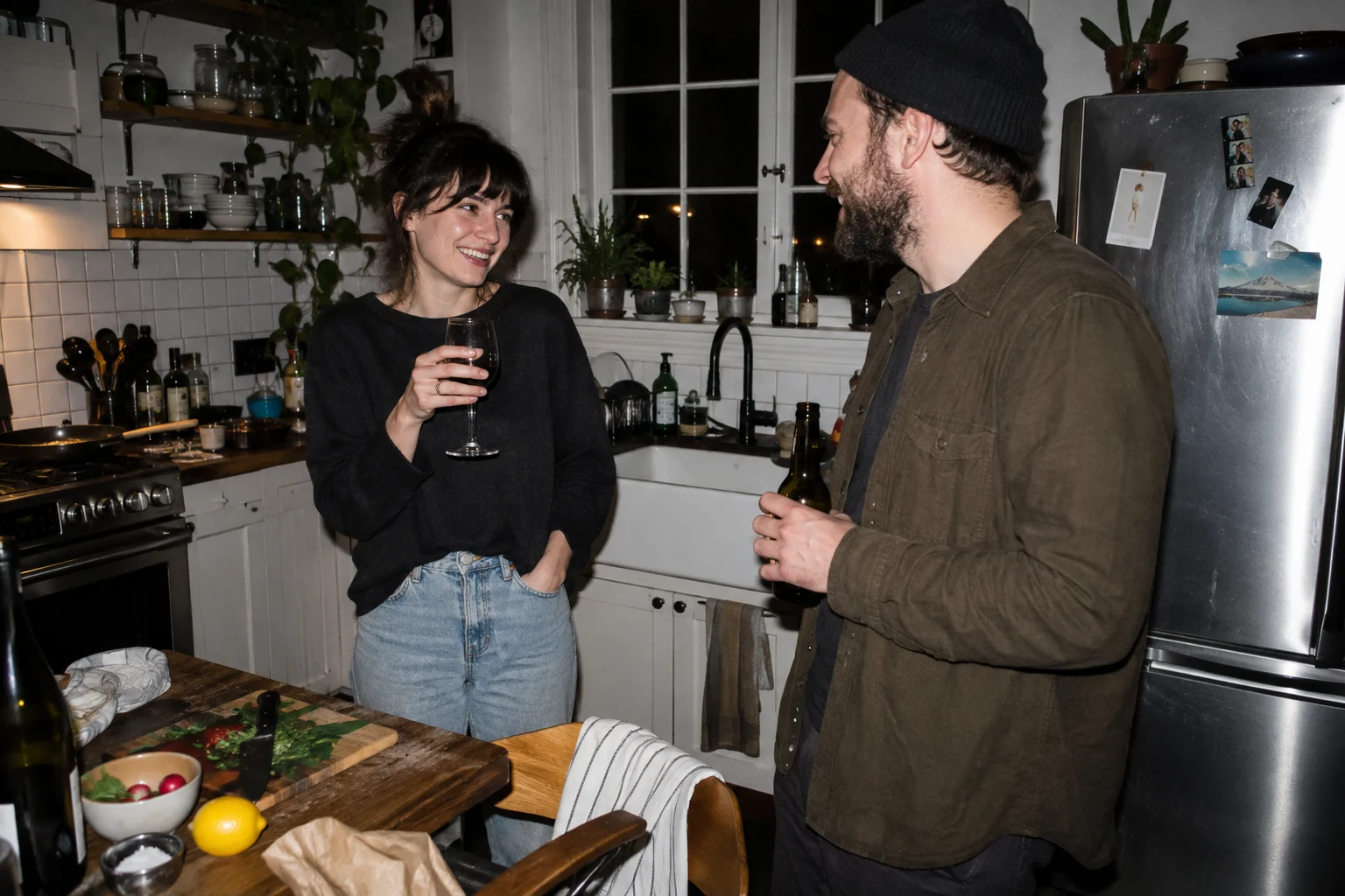 Two adult friends in casual clothes chatting in a cluttered home kitchen at night, hard on-camera direct flash, candid crop, natural faces.