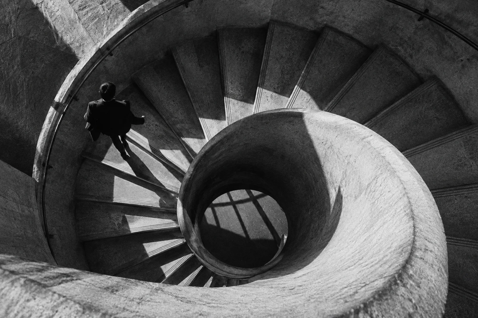 Overhead view of concrete spiral staircase and a single walking adult silhouette, hard side light and deep geometric shadows; monochrome.