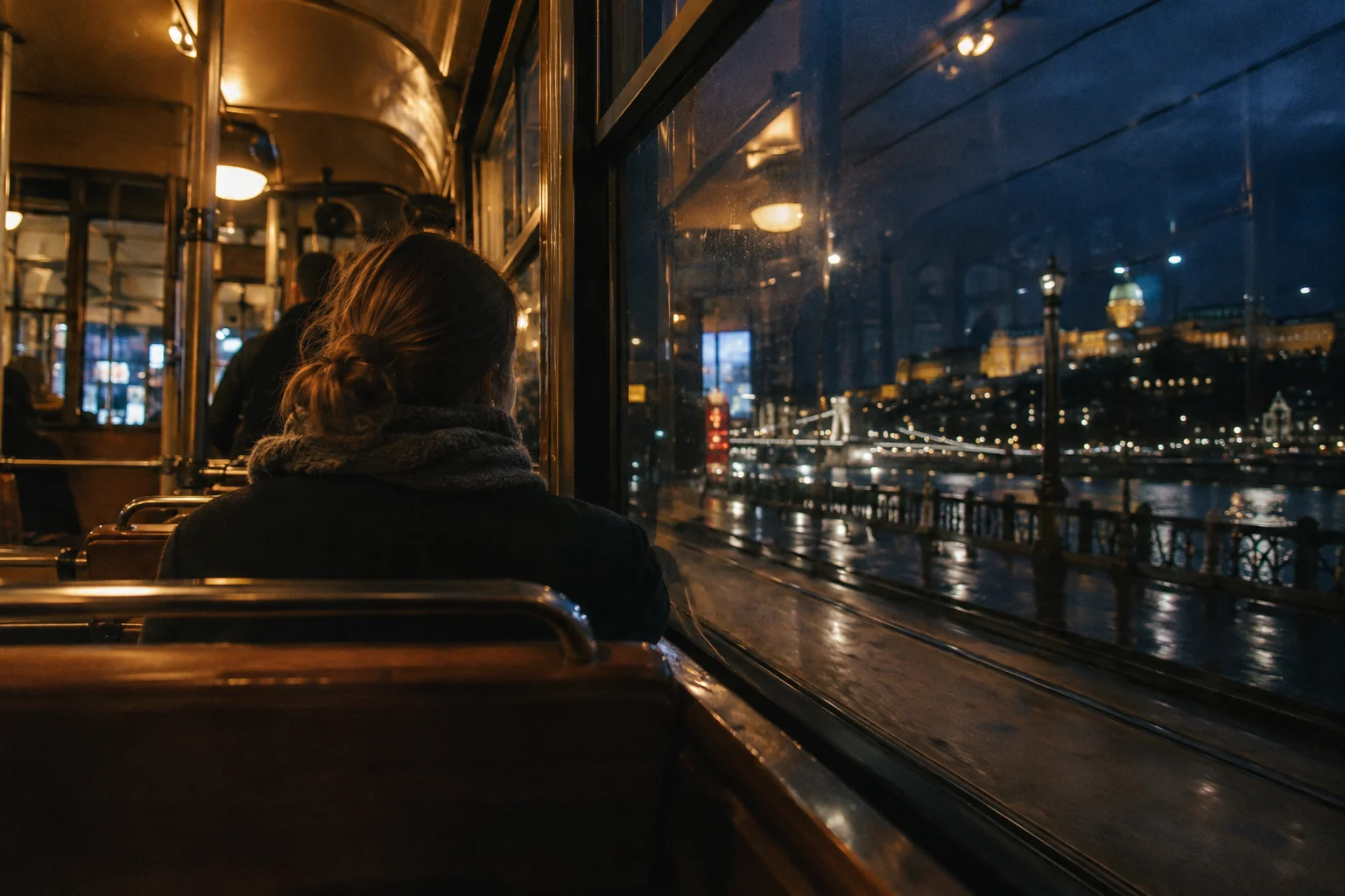 Adult passenger seen from behind inside a dim tram at night, warm carriage lamps and blue city outside, slight window reflections, available light film photograph no flash.