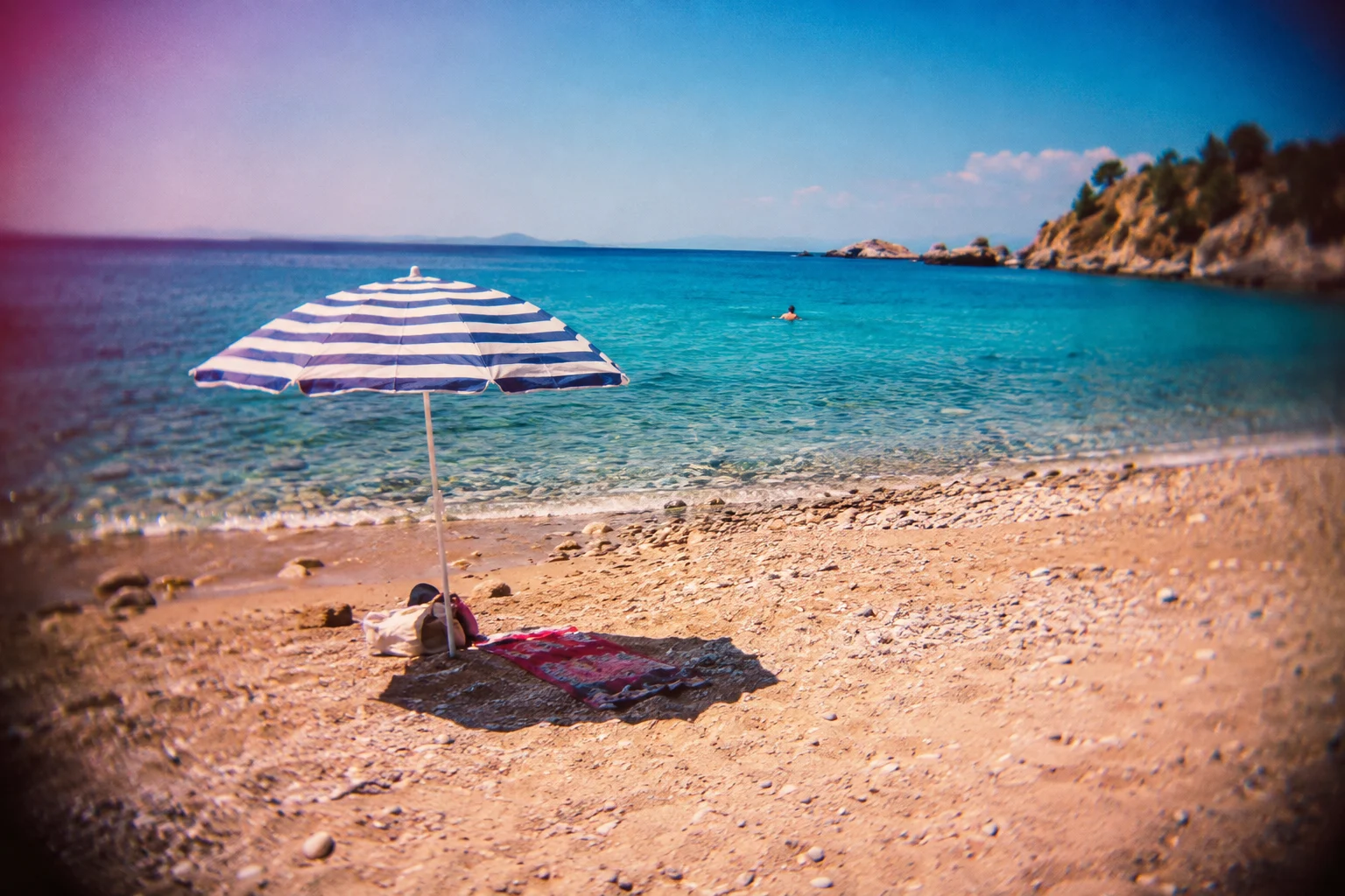 Empty beach with a striped parasol and a tiny distant adult swimmer; cheap plastic lens photograph, saturated cyan sea, magenta warm highlights, imperfect soft corners and optical vignette.