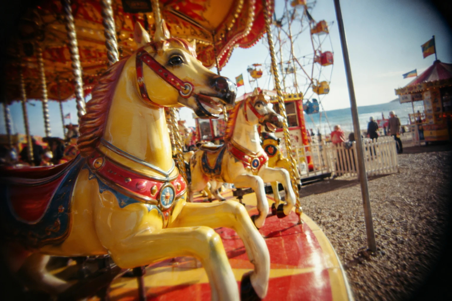 Small seaside amusement fair with colorful carousel horses in daylight, toy plastic lens snapshot, rich red and yellow, soft corners, dark vignette, slightly off-level framing.