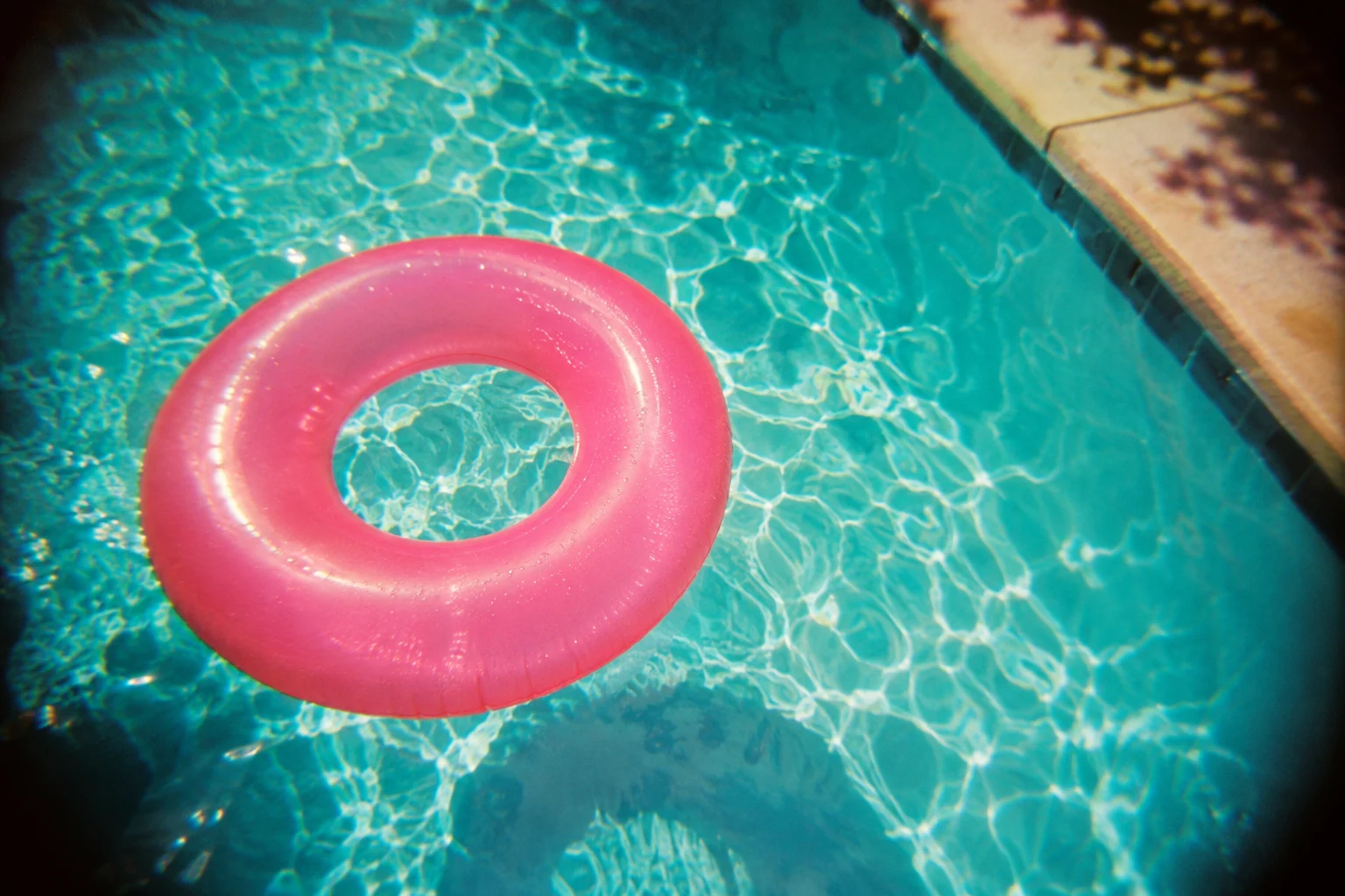 Pink inflatable ring floating in turquoise outdoor pool, top-down casual summer snapshot through toy camera plastic lens, soft edges and subtle color shift.